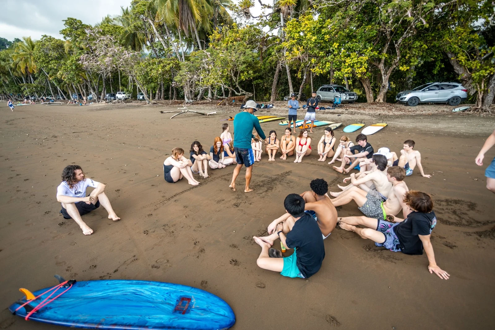 A Flow instructor briefing a group seated on the beach, with surfboards nearby